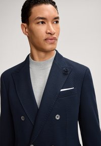 Marineblauwe dubbelgeknoopte blazer met visgraatstructuur, voorzien van een bloemaccent op het revers en een wit pochet. Daaronder een grijze coltrui.