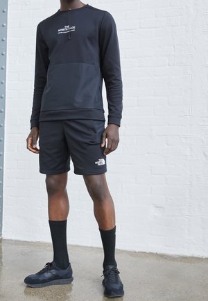 Mężczyzna w czarnej bluzie The North Face i szortach, czarne skarpety i czarne buty sportowe, stoi w pomieszczeniu na betonowej podłodze.