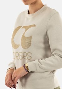 Sudadera de color beige claro con un logo en forma de "C" y texto en dorado brillante. Tiene un cuello redondo y puños acanalados, fabricada con un tejido suave.