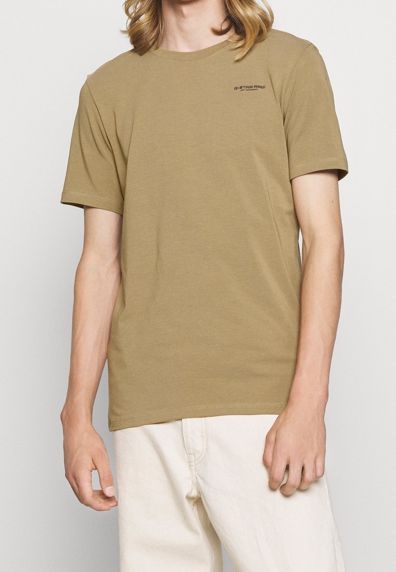 Personne portant un t-shirt kaki uni à manches courtes avec un petit logo noir "G-STAR RAW" sur la poitrine, associé à un pantalon blanc cassé.