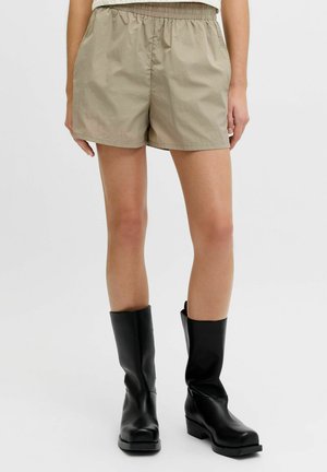 Oseba v bež elastičnih kratkih hlačah z visokim pasom in črnih usnjenih škornjih do sredine meč, stoji ob enostavni svetli podlagi.