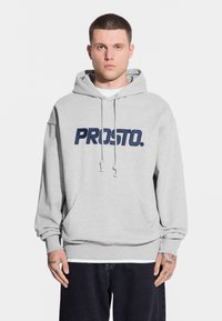 Jasnoszara bluza z kapturem z kieszenią kangurką, z dużym granatowym logo "PROSTO." na przodzie oraz sznurkiem do regulacji przy kapturze.