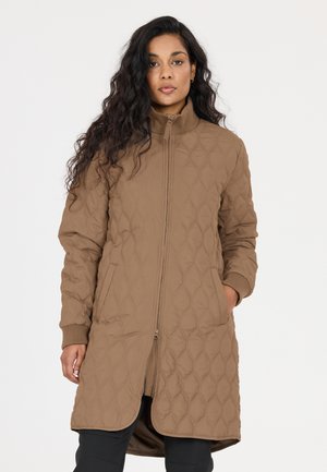 Woman stands facing forward, wearing a long, quilted brown coat with hands in pockets, against a plain light background.