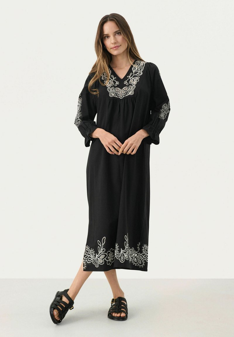 Schwarzes Kleid mit weißer Blumenstickerei am Ausschnitt und Saum, mit langen Ärmeln und lockerer Passform. Getragen mit schwarzen Sandalen.