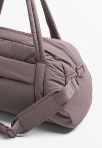 Sac de sport doux et rembourré en tissu mauve doux. Dispose de poignées robustes et d'une bandoulière ajustable. Texture lisse avec des coutures renforcées.