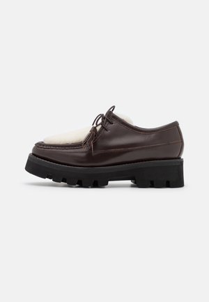 Chaussure à lacets en cuir marron foncé avec une semelle noire épaisse et un panneau en polaire crème sur le dessus, vue de côté sur fond blanc.