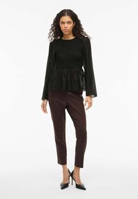 Top nero peplum con corpetto testurizzato, maniche lunghe in tessuto trasparente; abbinato a pantaloni aderenti marrone scuro e tacchi a spillo neri alti.