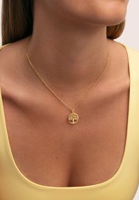Collier doré avec un pendentif circulaire arborant un motif d'arbre, reposant sur un cou lisse et dénudé sous un haut jaune.