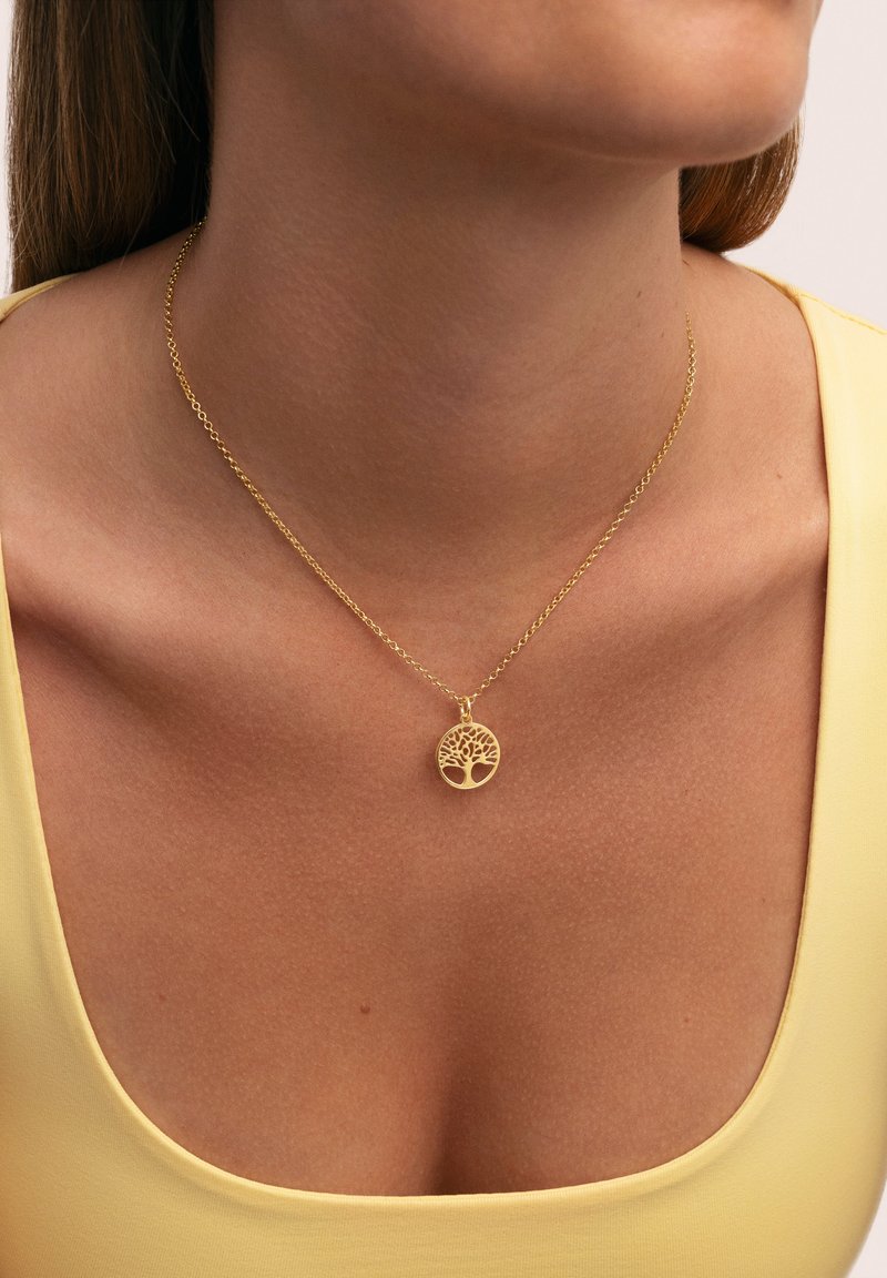 Collier doré avec un pendentif circulaire arborant un motif d'arbre, reposant sur un cou lisse et dénudé sous un haut jaune.