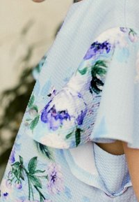 Blumengemustertes Kleid aus lichtblauem Stoff mit strukturierten Rüschen, in Farbtönen von Lila und Grün, und mit einem sanften Ausschnitt-Detail.