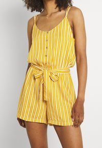 Žena v žlutém a bílém pruhovaném bezrukávovém overal s knoflíky vpředu a zavázaným páskem v pase.