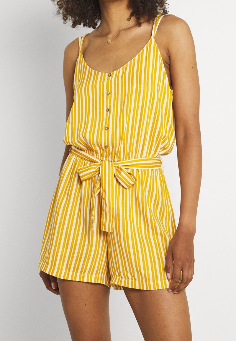 Žena v žlutém a bílém pruhovaném bezrukávovém overal s knoflíky vpředu a zavázaným páskem v pase.