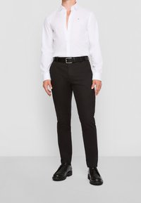 Camisa blanca de botón con un logo, cinturón negro y pantalones negros slim-fit. Zapatos negros con suelas texturizadas completan el conjunto.