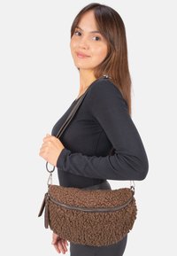 Sac à bandoulière texturé marron avec un extérieur duveteux, quincaillerie en argent et fermeture éclair. Sangle réglable attachée pour un port à l'épaule ou en travers du corps.