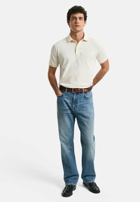 Beige polo skjorta med krage och korta ärmar, tillsammans med ljusblå raka jeans och svarta skor. Enkel design, avslappnad look.