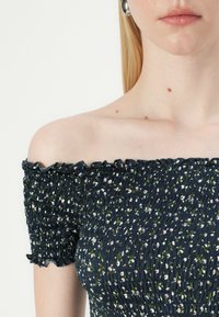 Bluza s spuščenimi rameni v temno modri barvi z cvetličnim vzorcem, z nabranimi detajli in nabrano robovi. Narejena iz lahkega materiala.