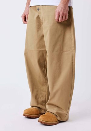 Personne portant un pantalon beige ample et des bottes en daim marron clair, debout devant un fond blanc uni.