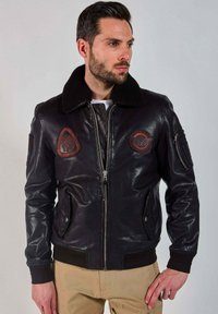 Homme portant une veste en cuir noir type bombardier avec des patches, un col en fourrure noire, une fermeture éclair à l'avant, et un pantalon beige, regardant vers la droite.