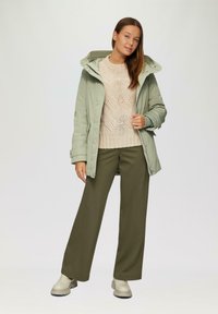 Veste imperméable vert clair avec capuche, pull en tricot beige, pantalon large olive et bottines crème, se tenant sur un fond uni.
