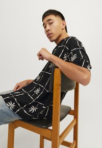 Camisa negra de manga corta con patrón de hojas de palma blancas y diseño en cuadrícula acolchada. Modelo sentado en una silla de madera, vistiendo jeans de color azul claro.