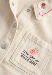 Koszula z kremowej, teksturowanej tkaniny, z srebrnymi guzikami, haftowanym kołnierzykiem z kwiatami oraz dekoracyjną kieszenią z motywem truskawki.