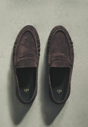 Mocassins en daim brun foncé avec un bout arrondi, des détails cousus et une texture douce. Intérieur noir avec un logo doré. Semelle plate.