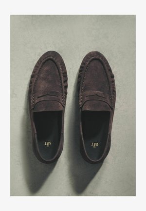 Mocasines de ante marrón oscuro con puntera redondeada, detalles cosidos y una textura suave. Interior negro con marca dorada. Suela plana.