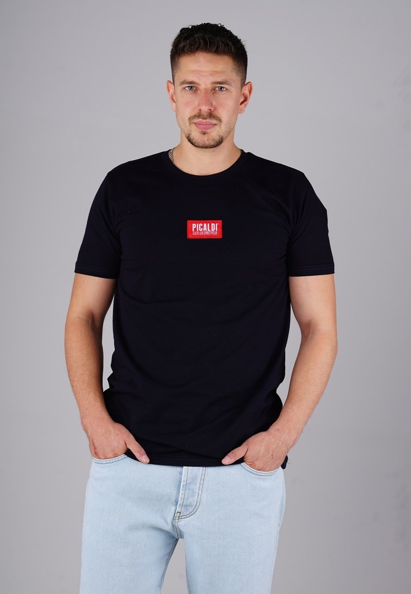 Mann mit kurzen Haaren und Bart, der ein schwarzes T-Shirt mit rotem Logo und hellblauen Jeans trägt, steht mit den Händen in den Taschen.