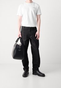 Uomo che indossa una t-shirt bianca Calvin Klein, jeans neri, stivali in pelle nera, mentre tiene una grande borsa da viaggio in pelle nera su uno sfondo bianco.
