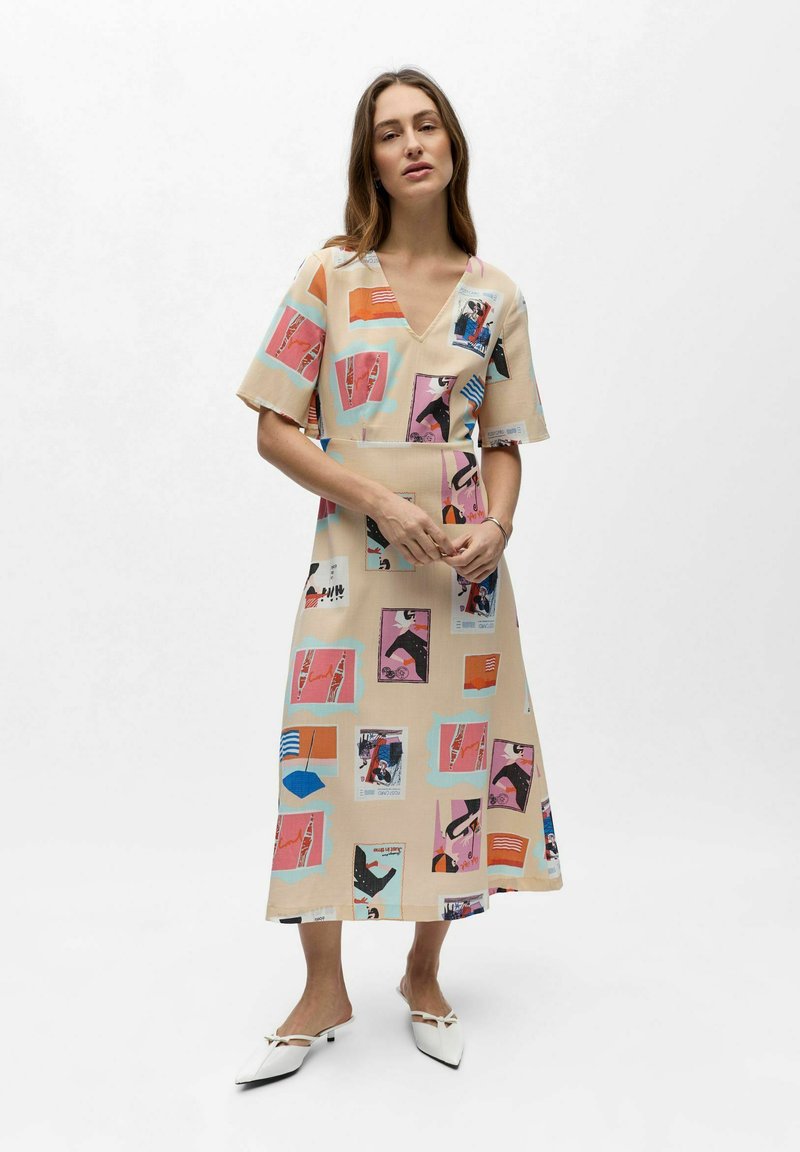 Femme portant une robe midi beige avec des imprimés abstraits et graphiques colorés, debout les mains jointes, sur un fond blanc uni.