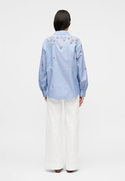 Γυναίκα με μακριά σκούρα μαλλιά φοράει μπλε ριγέ μπλούζα με λουλουδένιες εφαρμογές, λευκό παντελόνι με φαρδιά πόδια και καφέ παπούτσια, όρθια με την πλάτη προς τα εμπρός.