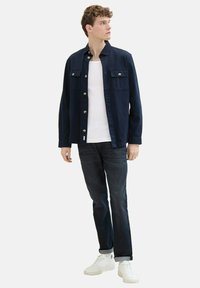 Chemise à boutons navy en coton avec deux poches à l'avant, portée sur un débardeur blanc, associée à un jean foncé et des baskets blanches.