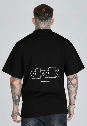 Homme avec des tatouages au cou et aux bras portant un t-shirt noir oversize avec les inscriptions blanches "siksilk" et "Have a nice day" dans le dos.