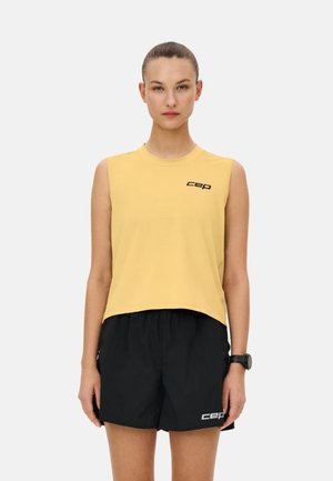 Woman with tied-back hair wearing a sleeveless yellow top and black shorts, standing with arms relaxed against a plain background.