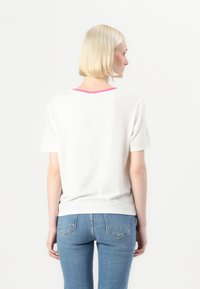 T-shirt branca de manga curta com detalhe em colarinho rosa, feita de material suave, apresentada por um modelo de costas, combinada com jeans azuis.