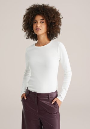 Femme aux cheveux bouclés portant des boucles d'oreilles en or, un haut à manches longues blanc et un pantalon en cuir bordeaux taille haute, les mains dans les poches.