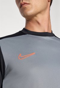 Nike treningsoverdel i grått med svarte detaljer. Har en tettsittende design, glatt tekstur og fremtredende oransje swoosh-logo.