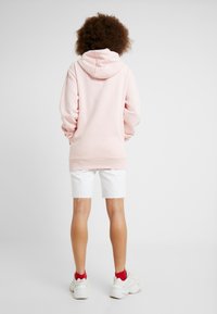 Sudadera rosa con capucha y mangas largas, hecha de tejido suave. Llevada con pantalones cortos blancos. Las zapatillas blancas tienen acentos rojos.