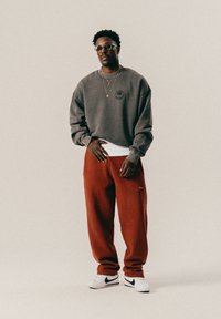 Άνδρας που φοράει γυαλιά, γκρι φούτερ, κόκκινες φόρμες, λευκό πουκάμισο με στρώσεις και λευκά αθλητικά παπούτσια Nike, στέκεται σε απλό φόντο.