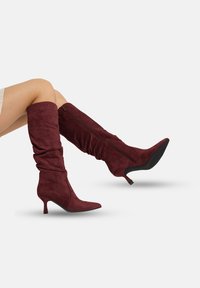 Bordeauxrode laarzen tot aan de knie, gemaakt van zacht, gestructureerd materiaal met een puntige neus, een kleine hak en een ritssluiting aan de zijkant. Ontwerp met een nonchalante pasvorm.