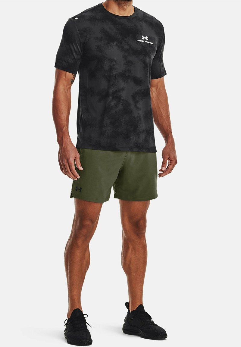 Man wearing black camo-patterned Under Armour t-shirt, olive green shorts, and black athletic shoes, standing against white background.