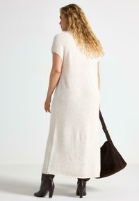 Kvinde med langt krøllet hår iført en hvid struktureret midikjole, mørkebrune hæler og bærende på en stor mørkebrun sømhåndtaske.