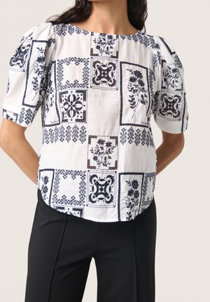 Vrouw draagt een witte blouse met zwarte bloemen- en geometrische vierkante patronen en pofmouwen, gecombineerd met een zwarte broek.