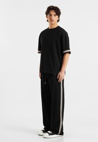 Svart kortermet t-skjorte med kontrasterende mønsterkant på ermene, sammen med svarte joggebukser med sidestriper, og svarte og hvite sneakers.