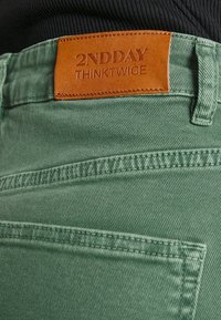 Pantalones de mezclilla verde con una superficie texturizada, que cuentan con una etiqueta de cuero marrón con la inscripción "2NDDAY THINKTWICE" grabada en la cintura.