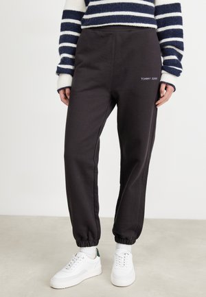 Osoba ubrana w czarne spodnie dresowe Tommy Jeans, biało-granatowy sweter w paski, białe skarpetki i białe trampki, stojąca na jasnej podłodze.