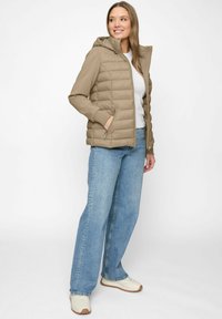 Frau in beiger Daunenjacke mit Kapuze, weißem T-Shirt, weiten blauen Jeans und weißen Sneakers, die vor einem weißen Hintergrund steht.