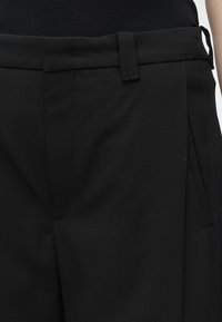 Gros plan sur un pantalon noir tailleur avec passants pour ceinture, une fermeture éclair cachée à l'avant et des poches latérales, porté avec un haut noir.