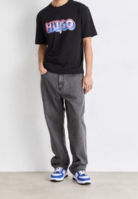 Svart kortärmad t-shirt med färggrafik "HUGO", grå avslappnade jeans och blå och vita sneakers med svart sula.