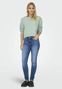 Pull en vert clair texturé avec des manches retroussées, associé à un jean bleu ajusté et des chaussures noires à enfiler, le mannequin est de face.